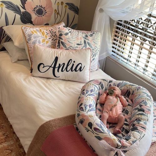 Baby in a floral bassinet with 'Anlia' pillow on a bed