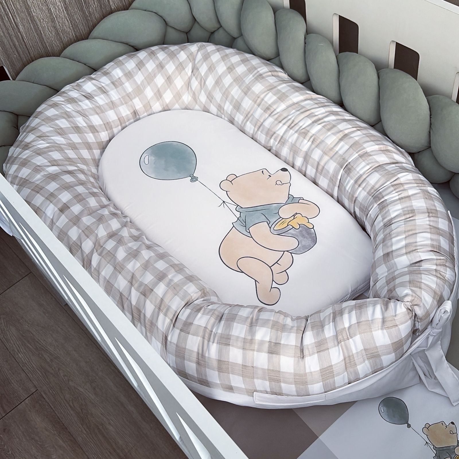 Baby nest with checkered border and Winnie the Pooh design inside, placed in a crib.