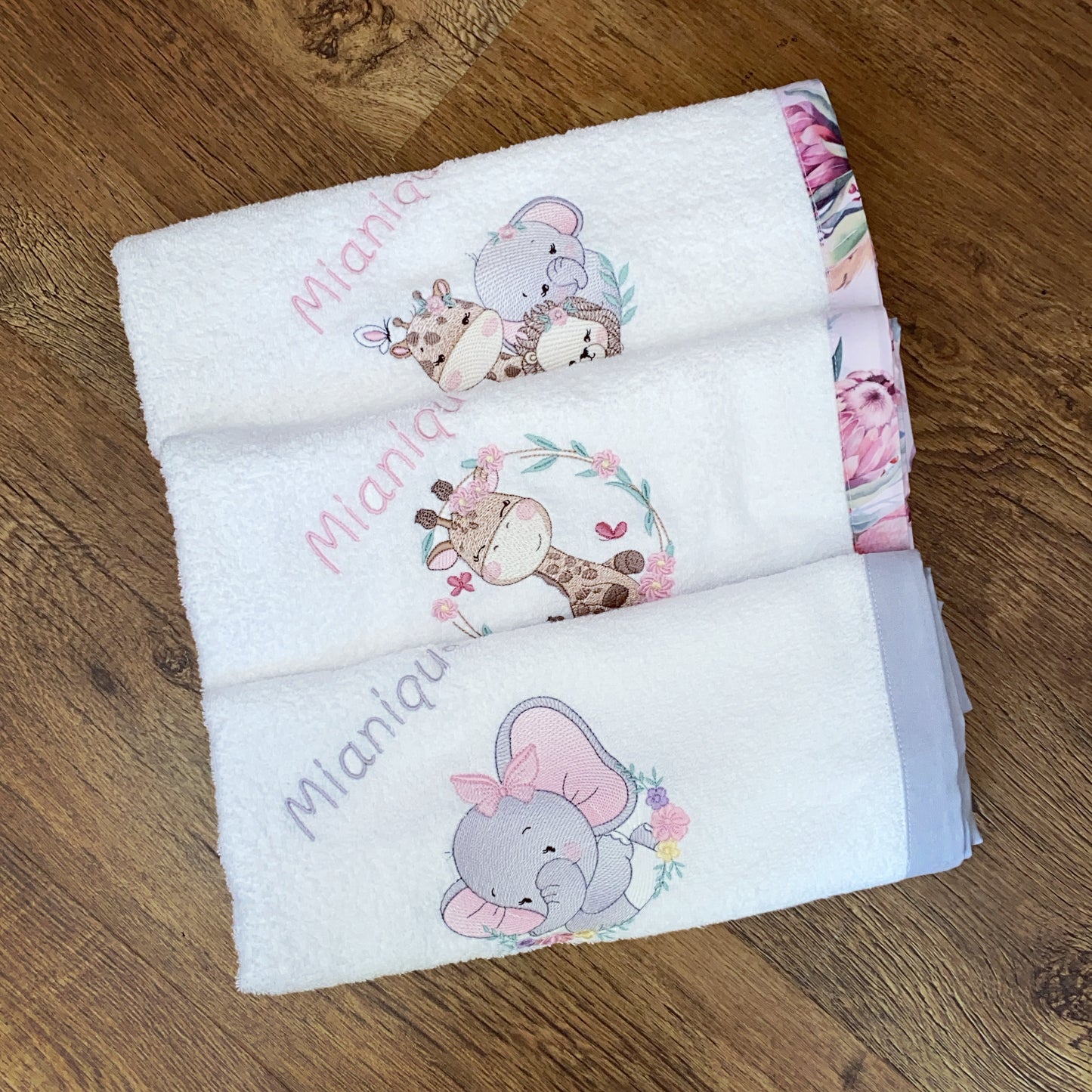 Stack of baby Burpcloths - Pink Safari 01 with animal designs and embroidered names on a wooden surface