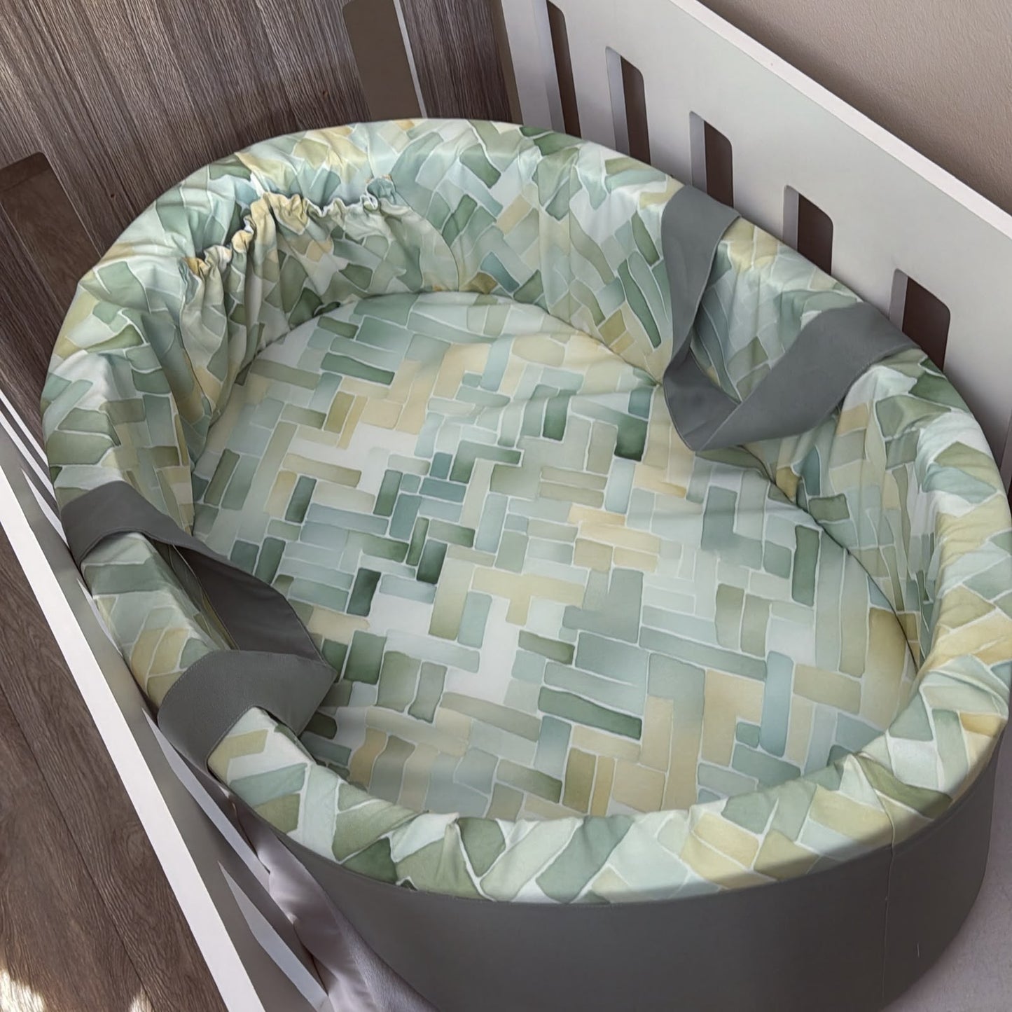 A round baby bassinet cushion set with a geometric patterned fabric in shades of sage green, yellow, and white, featuring gray straps, shown inside a white wooden bassinet.