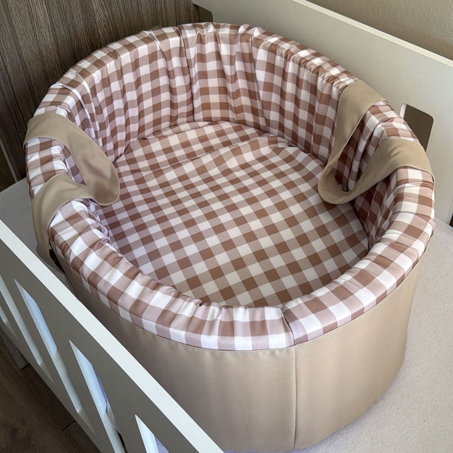 Carry cot with brown and white checkered pattern and beige trim on a wooden floor.