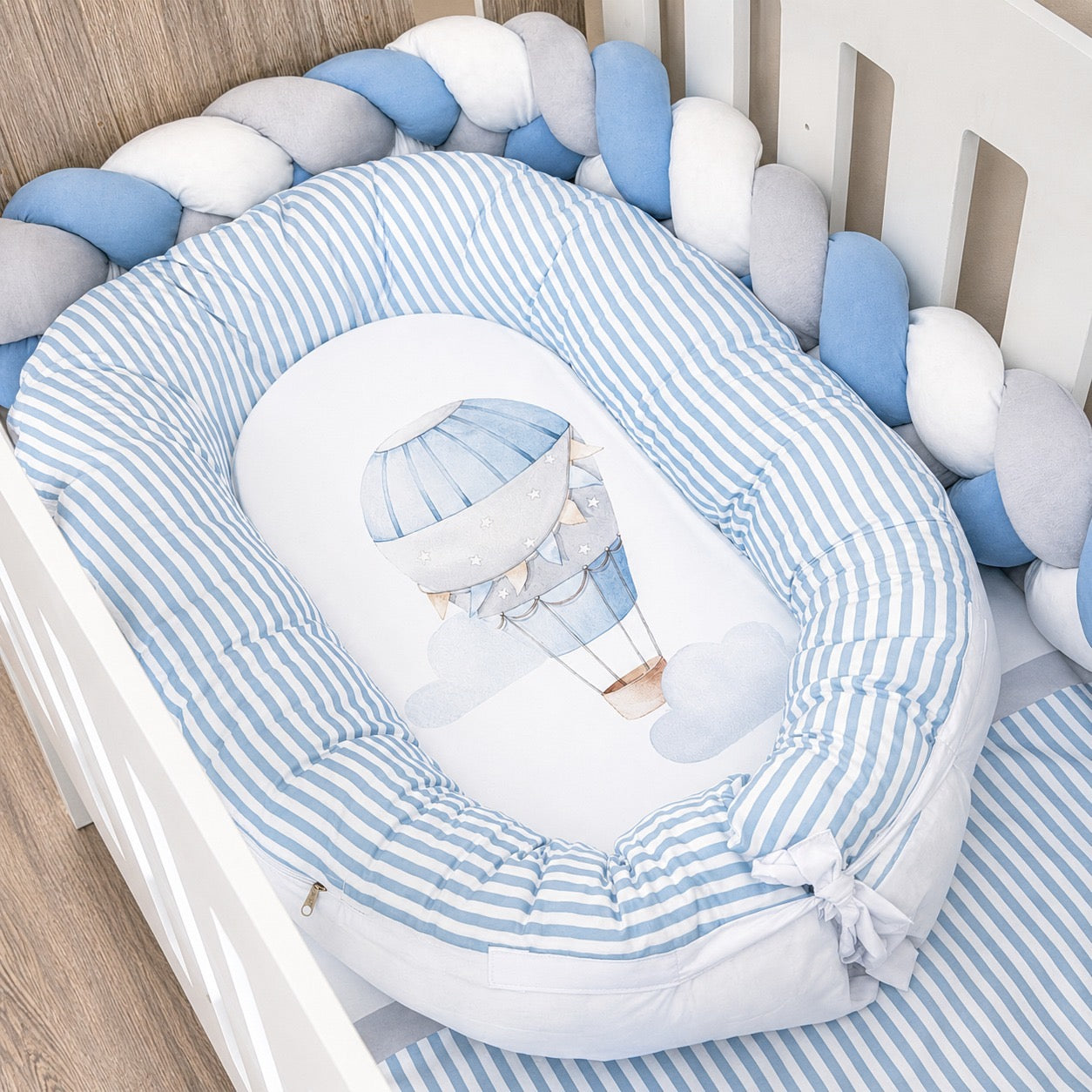 A baby sleep guard or bassinet cushion set in a white wooden crib, featuring a blue and white striped padded mattress with a hot air balloon illustration in the center, surrounded by a braided blue, white, and gray bumper pad.