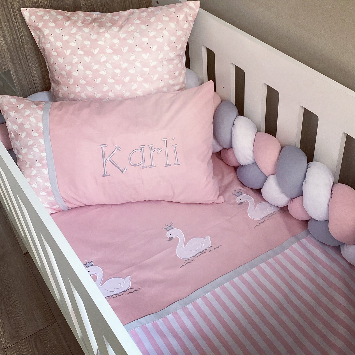 Crib with pink bedding featuring swan designs and a pillow with 'Karli' embroidered on it.