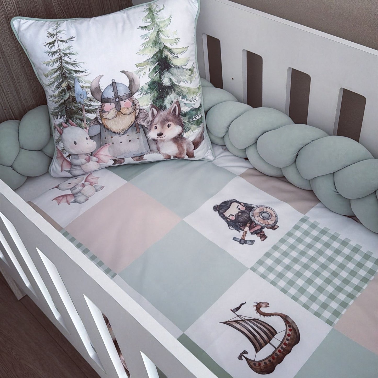 A baby crib bedding set featuring a patchwork quilt in sage green, beige, and white with Viking-themed illustrations, a matching braided bumper, and a decorative pillow with a Viking, wolf, and rabbit illustration.