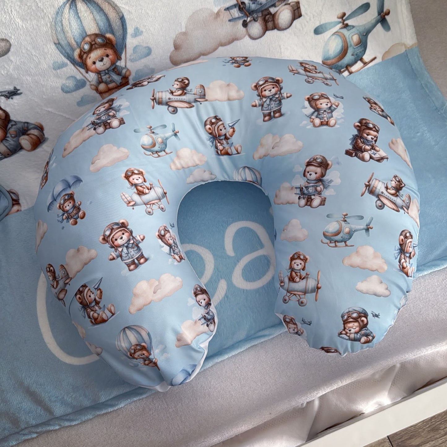 A light blue baby support and breastfeeding pillow with a cartoon teddy bear and airplane pattern, resting on a matching baby blanket.