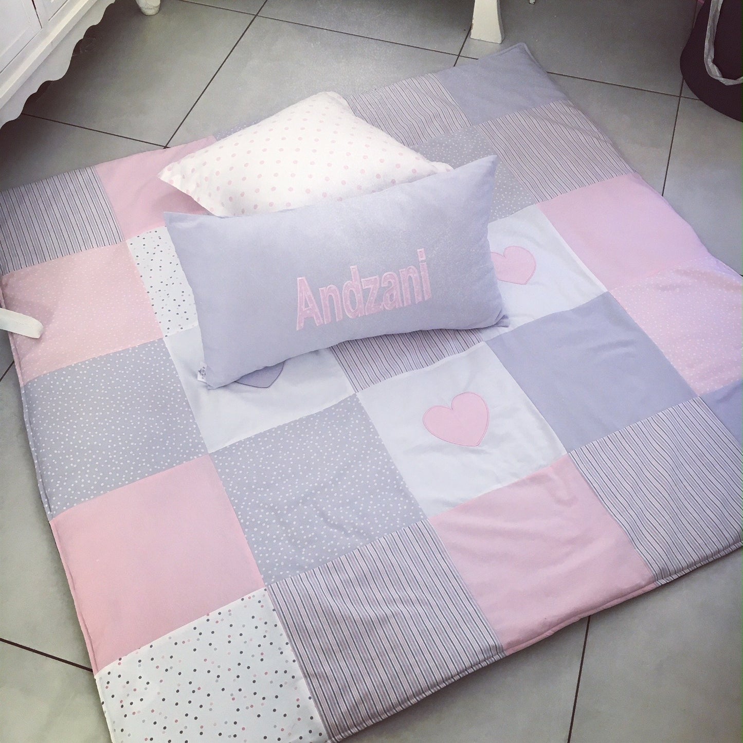 Patchwork blanket with pink and gray squares and a heart design, featuring a pillow with 'Andzani' on a tiled floor.