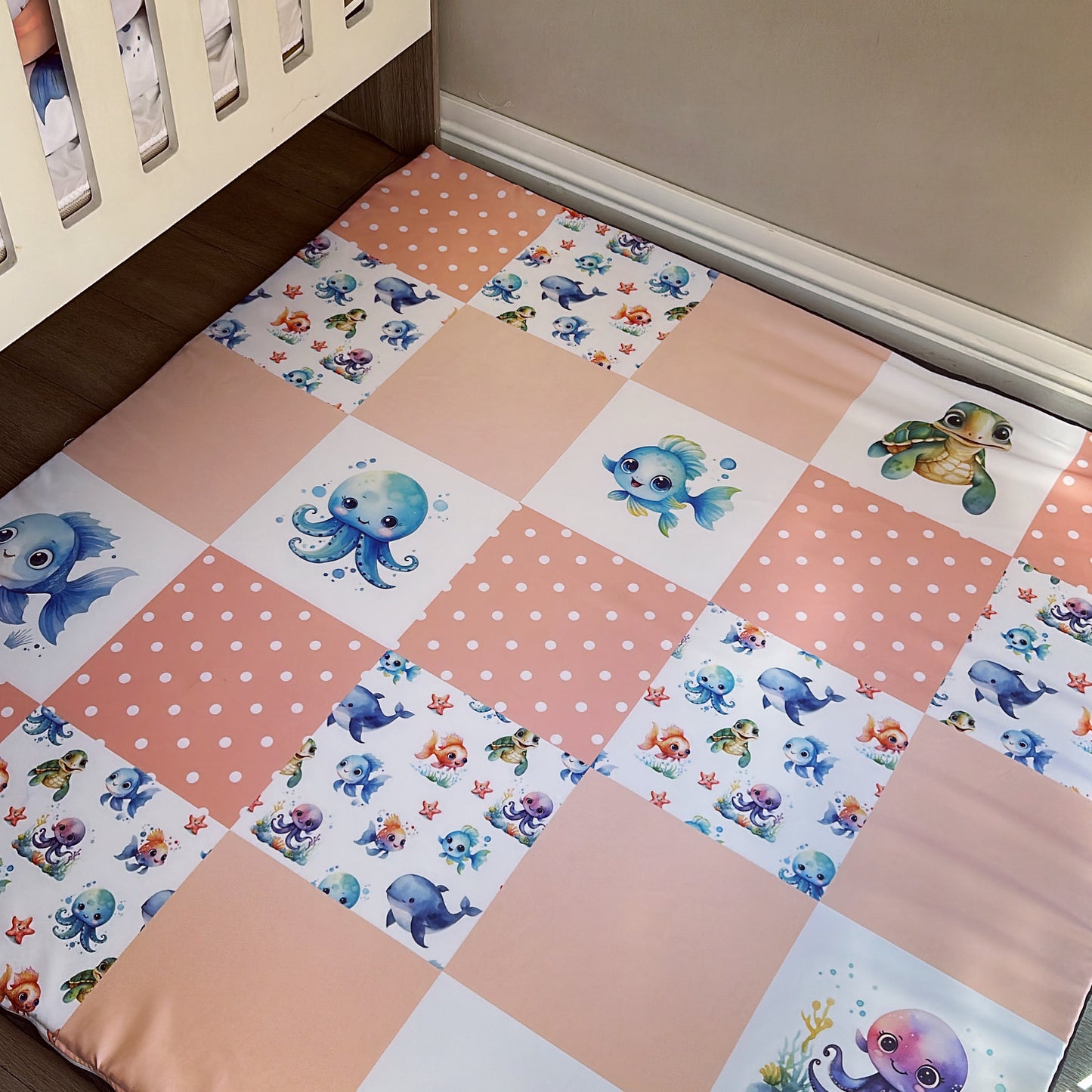 A large pink playmat with an ocean theme, featuring various sea creature illustrations, placed on the floor next to a baby's crib.