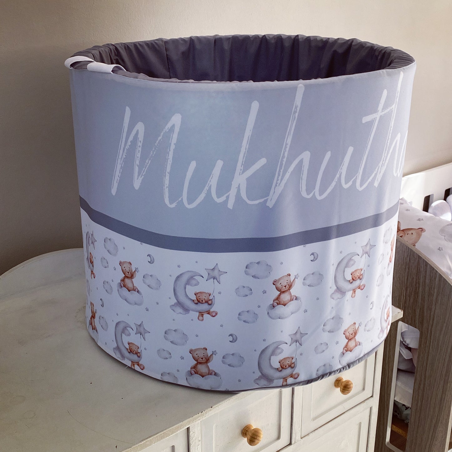 A large grey laundry barrel with a bear pattern and the text 'MUKHUTA' on it, placed on a wooden furniture item in a room with a light grey wall.