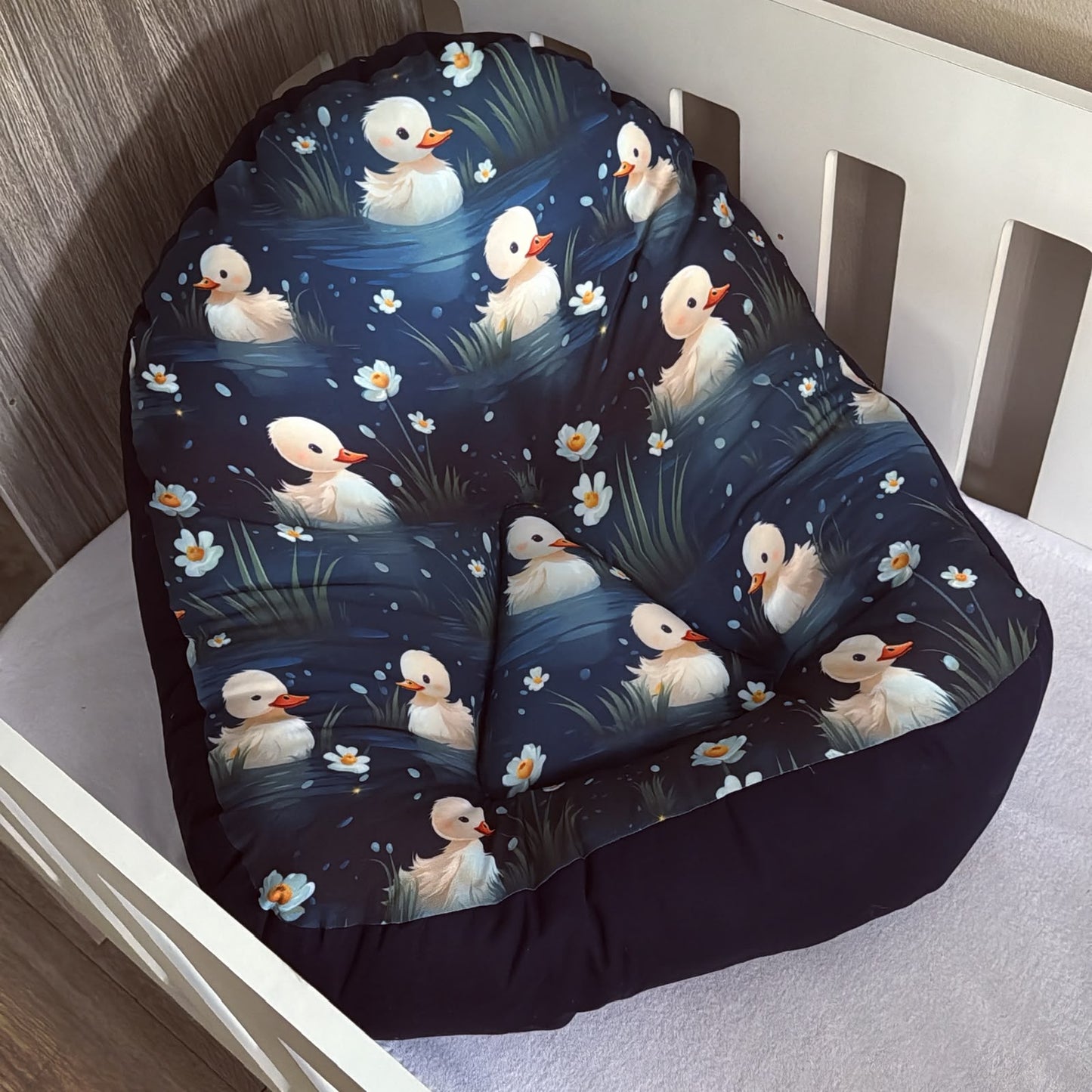 A plush bean bag chair featuring a printed pattern of white ducks in a blue pond with white flowers, placed on a white wooden bed frame.