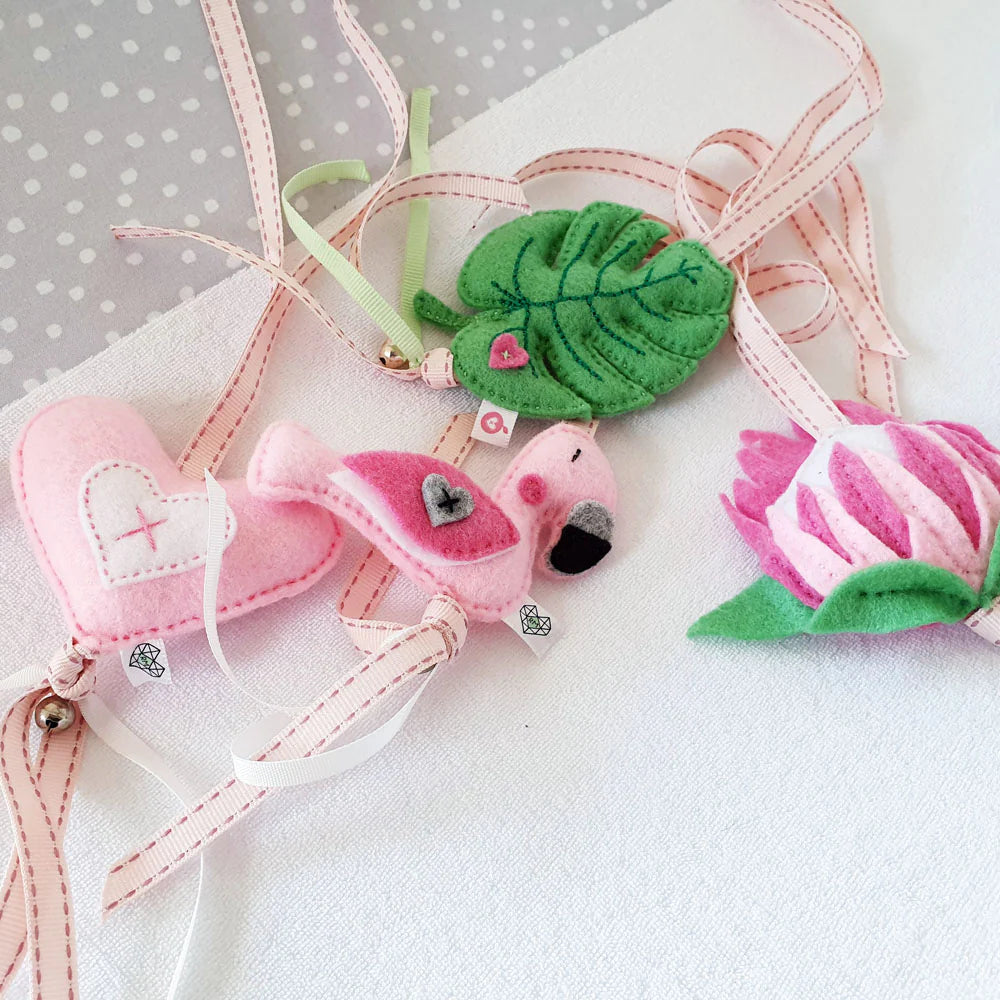A set of handmade felt bookmarks shaped like a pink heart, a pink flamingo, a green monstera leaf, and a pink-and-white striped flower, each attached to a pink-and-white ribbon.