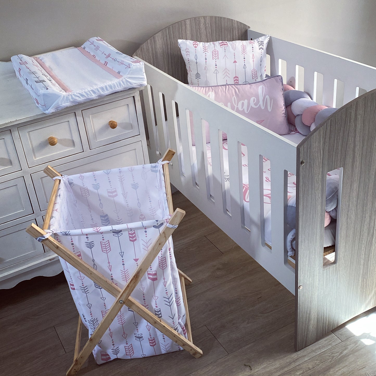 A wooden laundry basket with a white cushion and pink patterned fabric, placed in a nursery room setting with a wooden crib and a side table.