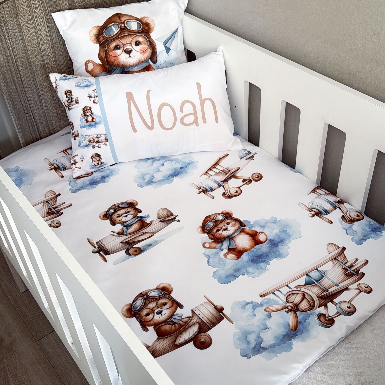 Children's crib with teddy bear and airplane patterned bedding, featuring a pillow with a teddy bear wearing goggles and the name 'Noah'.