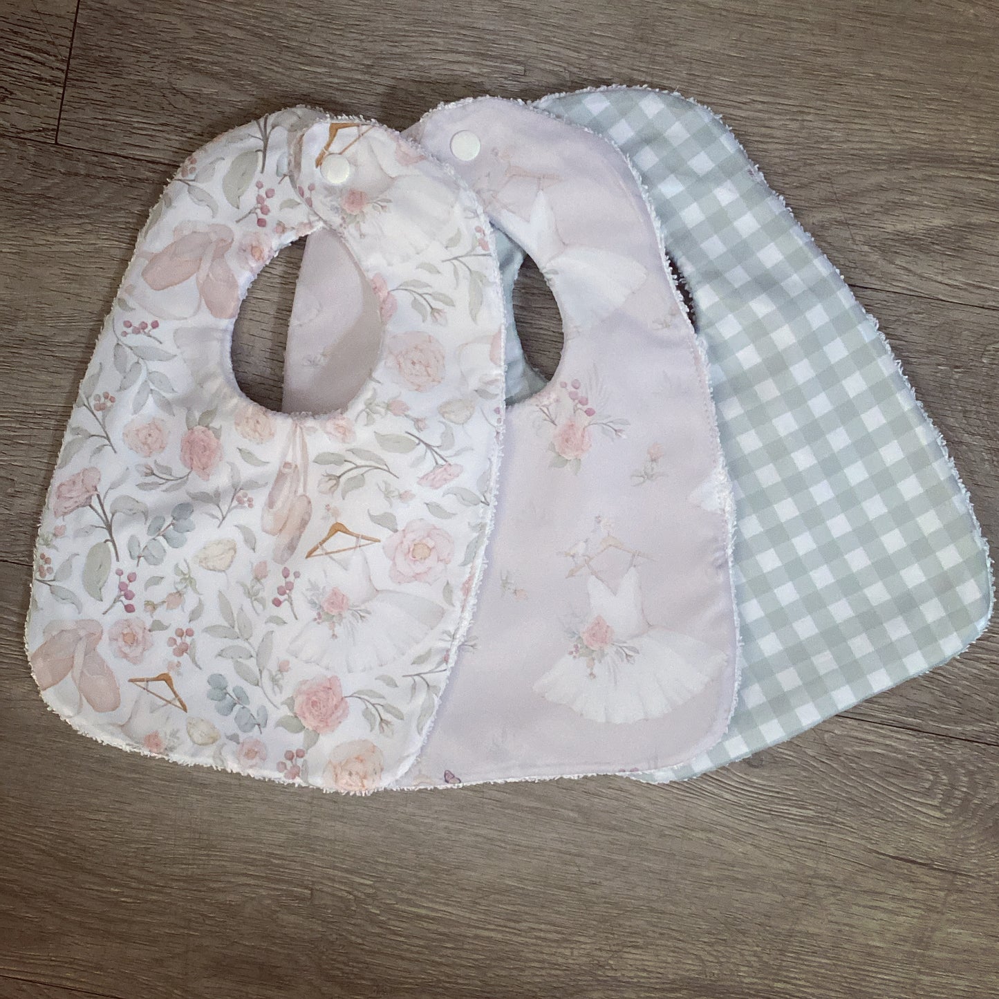 Three baby bibs with floral, checkered, and plain designs on a wooden surface.