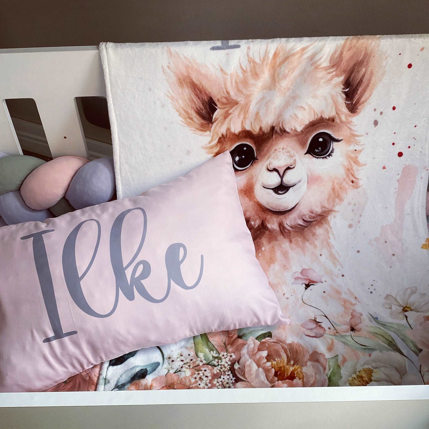 Floral blanket with a llama design and a pillow with 'Ilke' on a bed.