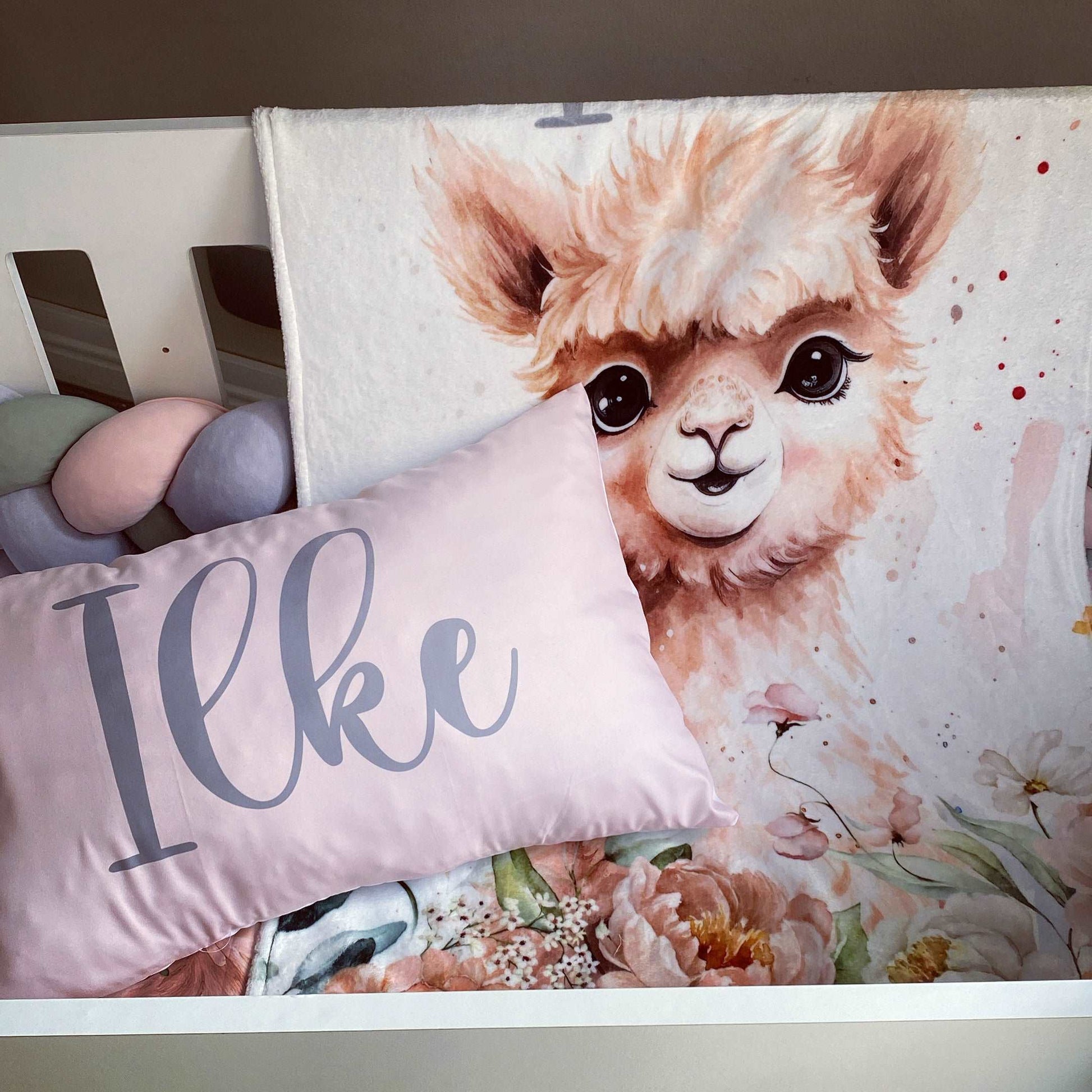 Floral blanket with a llama design and a pillow with 'Ilke' on a bed.