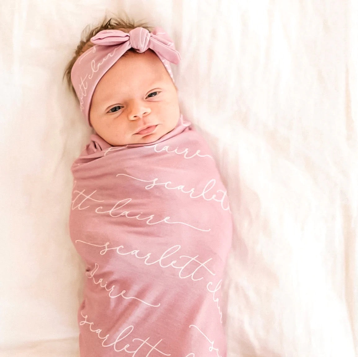 Baby wrapped in a pink blanket with text, wearing a matching headband, on a soft white background.