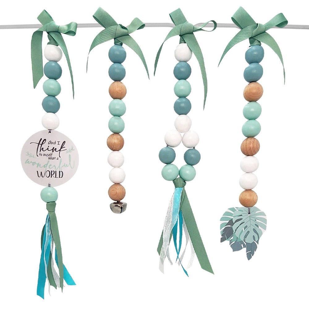 A decorative hanging garland with four vertical strands of wooden beads in mint green, white, and natural wood tones, tied to a white rod with mint green ribbons. One strand ends with a round white tag reading 'I think the world is a wonderful place,' another has white and mint beads with a small silver bell, and one ends with a monstera leaf charm.