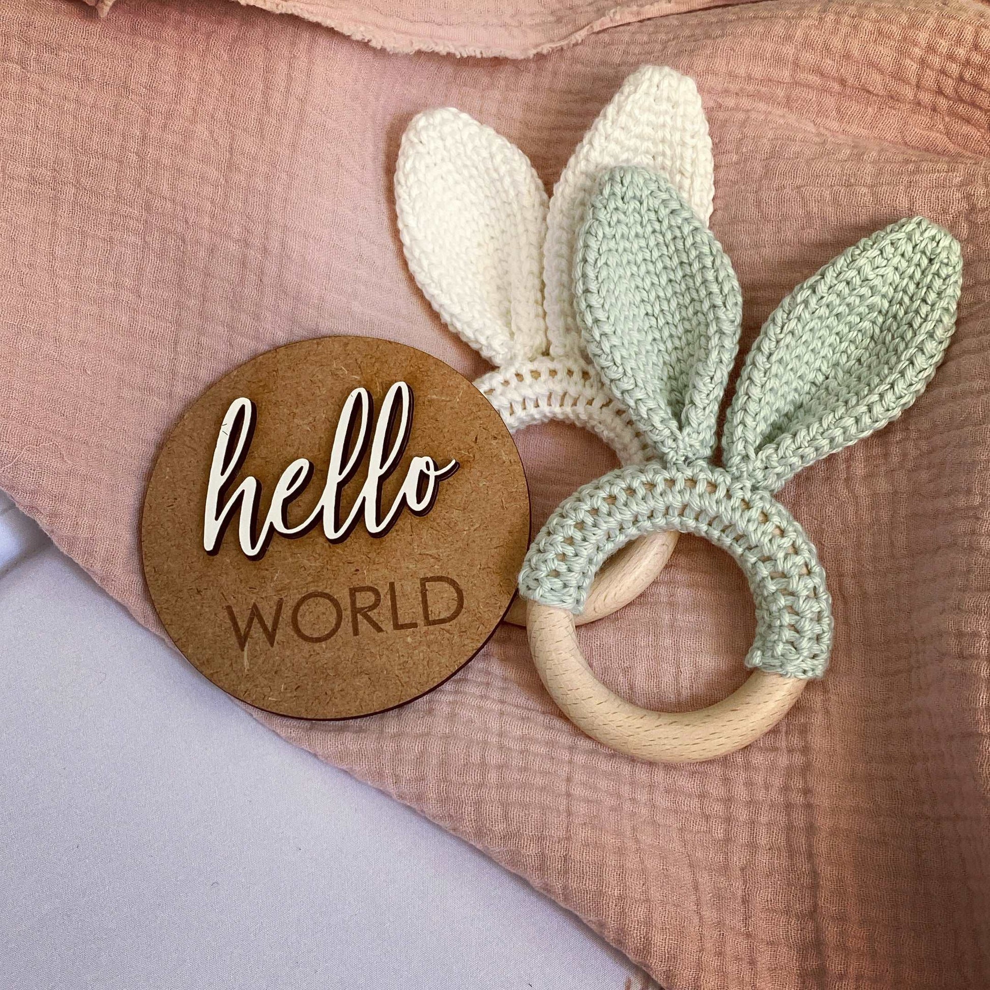 Crocheted bunny ear teething ring with 'hello world' wooden tag on a soft fabric background