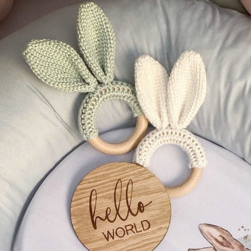 Two knitted bunny ear rings on a white surface with a wooden disc that says 'hello WORLD'.