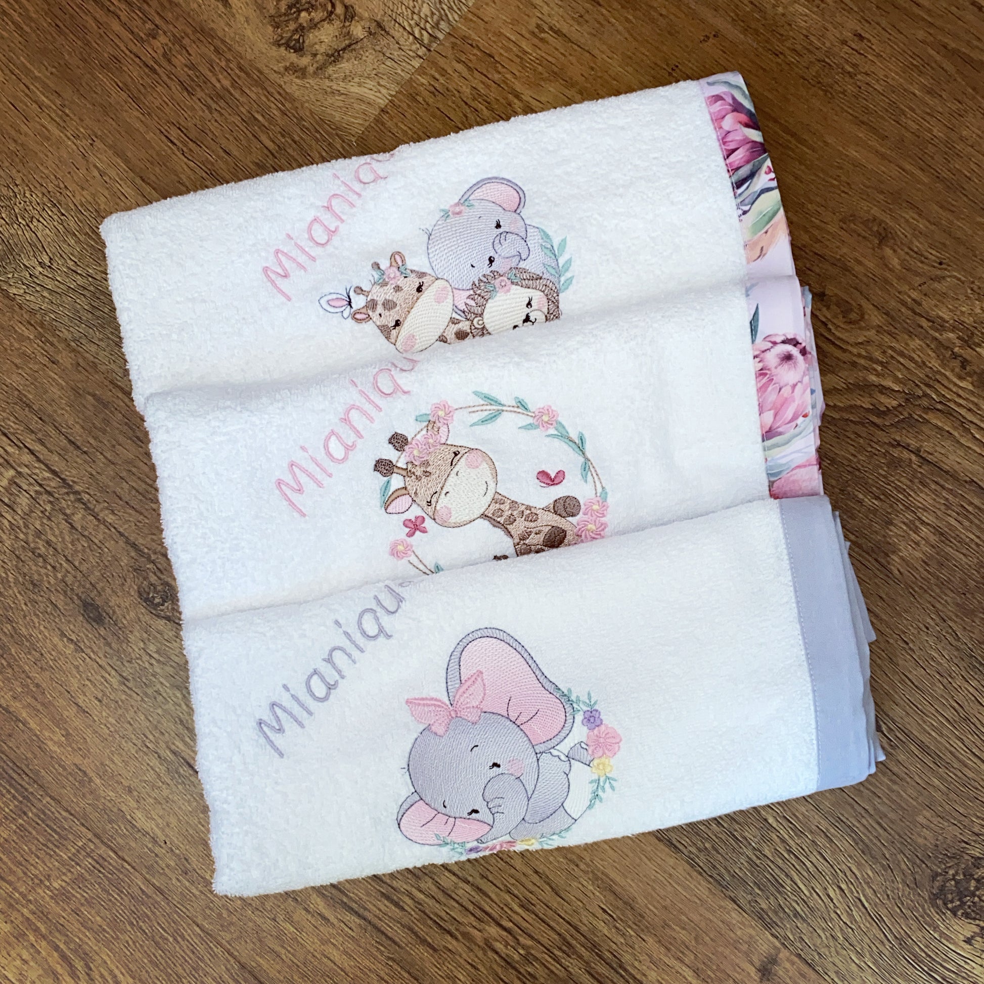 Stack of baby Burpcloths - Pink Safari 01 with animal designs and embroidered names on a wooden surface