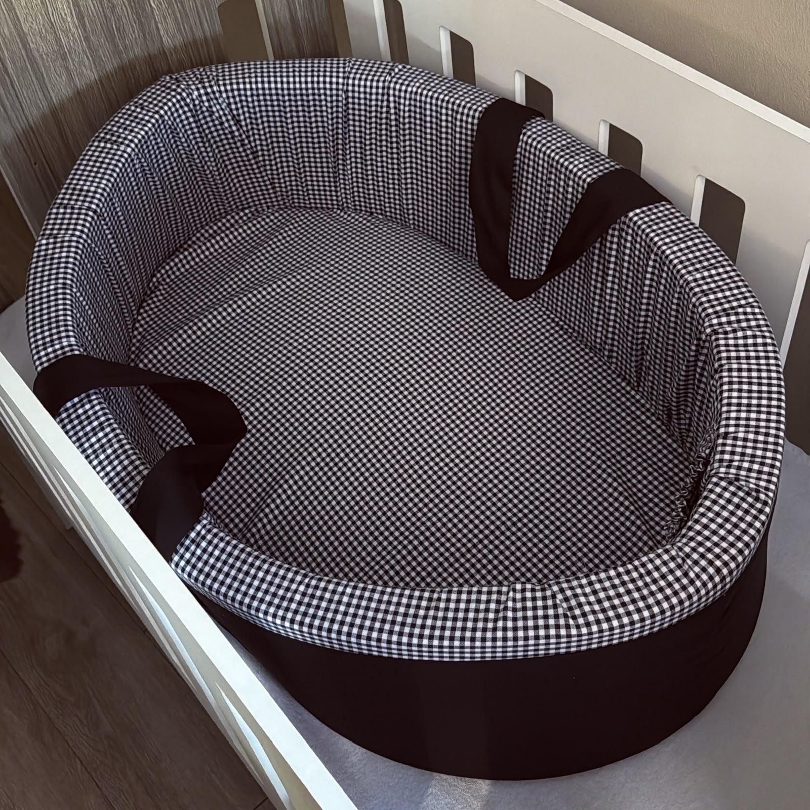 An oval carry cot with a black and white gingham check pattern and dark brown trim and handles, shown placed inside a white crib.