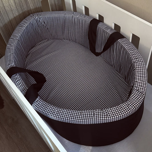 An oval carry cot with a black and white gingham check pattern and dark brown trim and handles, shown placed inside a white crib.