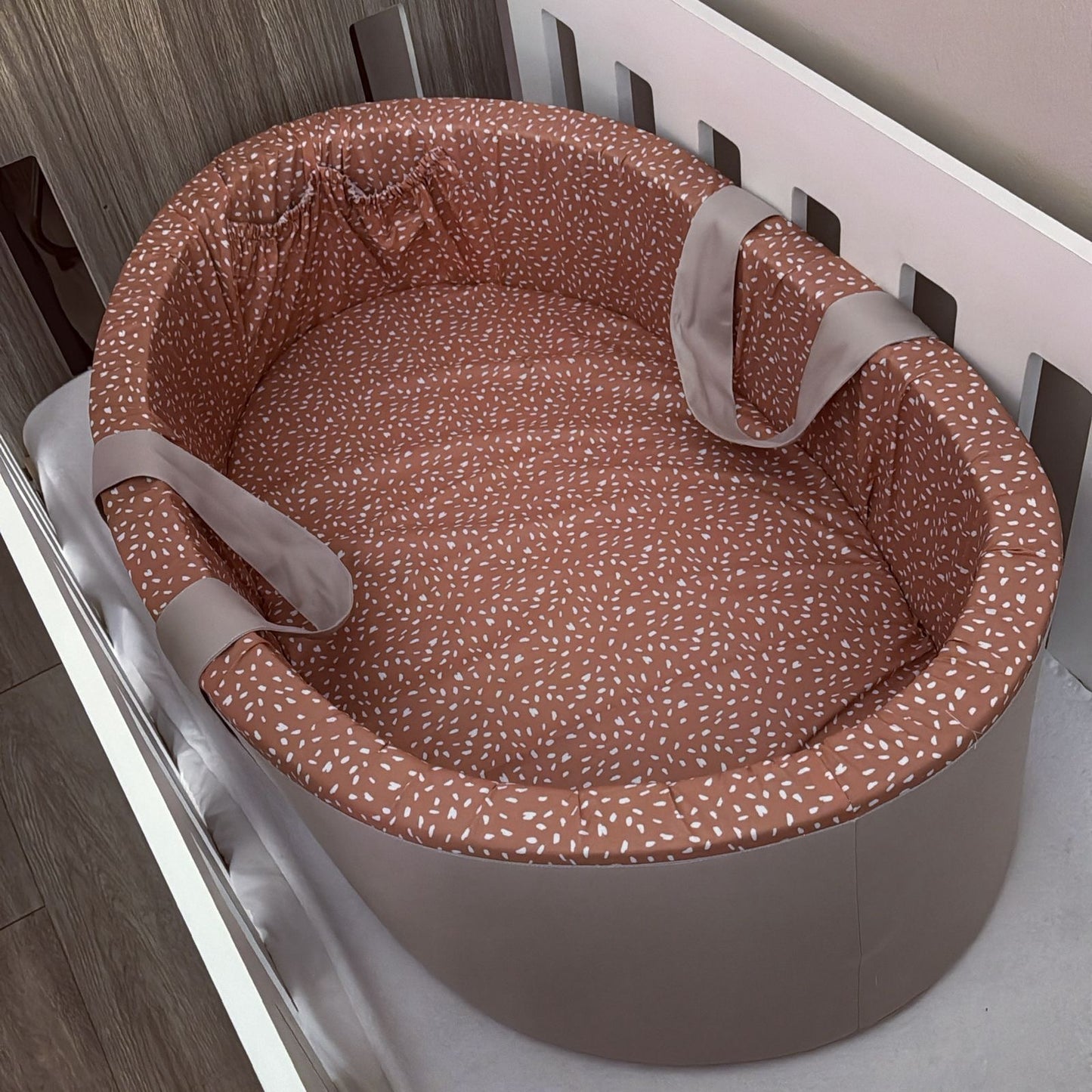 Baby carry cot bassinet with a terracotta rust-coloured fabric liner covered in small white speckles, featuring light gray safety straps, shown inside a white wooden crib.
