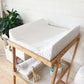 A white changing pad on a light wood changing table stand, styled in a bright nursery setting with a white paneled wall, a green plant, and baby accessories. | Baby Change Mats | Soft Spot Kids | South Africa
