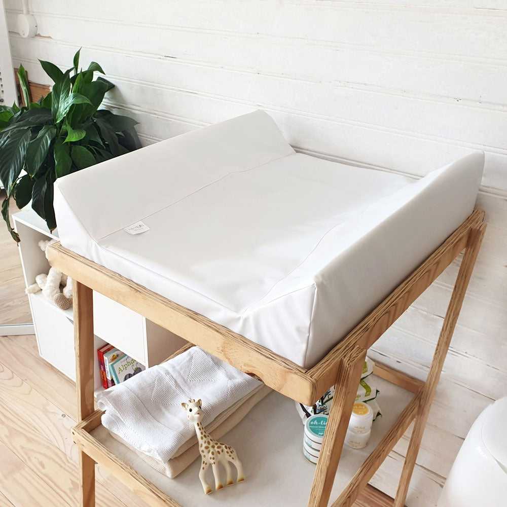 A white changing pad on a light wood changing table stand, styled in a bright nursery setting with a white paneled wall, a green plant, and baby accessories.