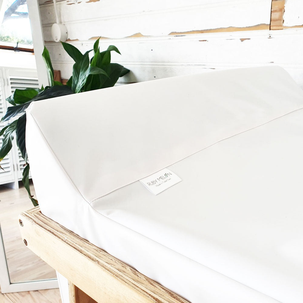 A white padded changing table pad on a wooden frame in a bright indoor room with white brick walls and a green plant in the background. A small sewn label is visible on the front.