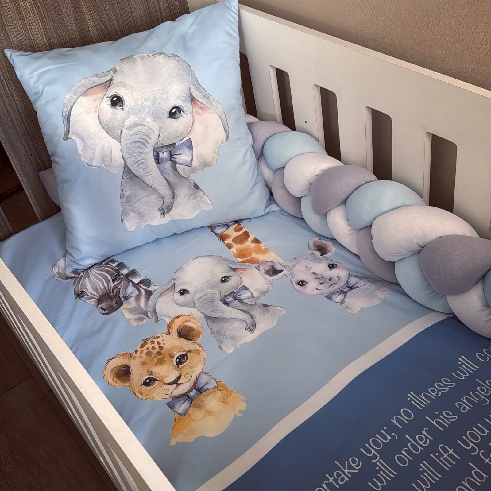 A baby bedding set in a white crib, featuring a light blue sheet and matching pillowcase printed with watercolor illustrations of baby animals including an elephant, zebra, giraffe, lion cub, and a pig. A chunky braided bumper in matching light blue, white, and gray is attached along the side.