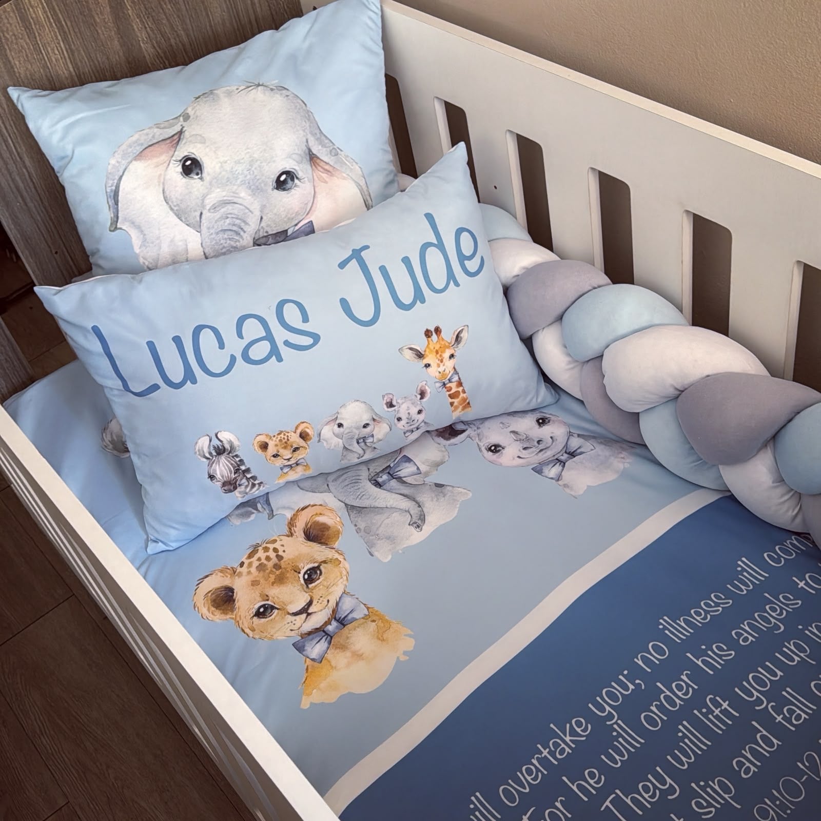 A baby bedding set styled inside a white wooden crib, featuring a light blue crib sheet, a matching pillow with animal illustrations, a personalized pillow reading 'Lucas Jude', and a braided bumper in shades of blue, gray, and white.