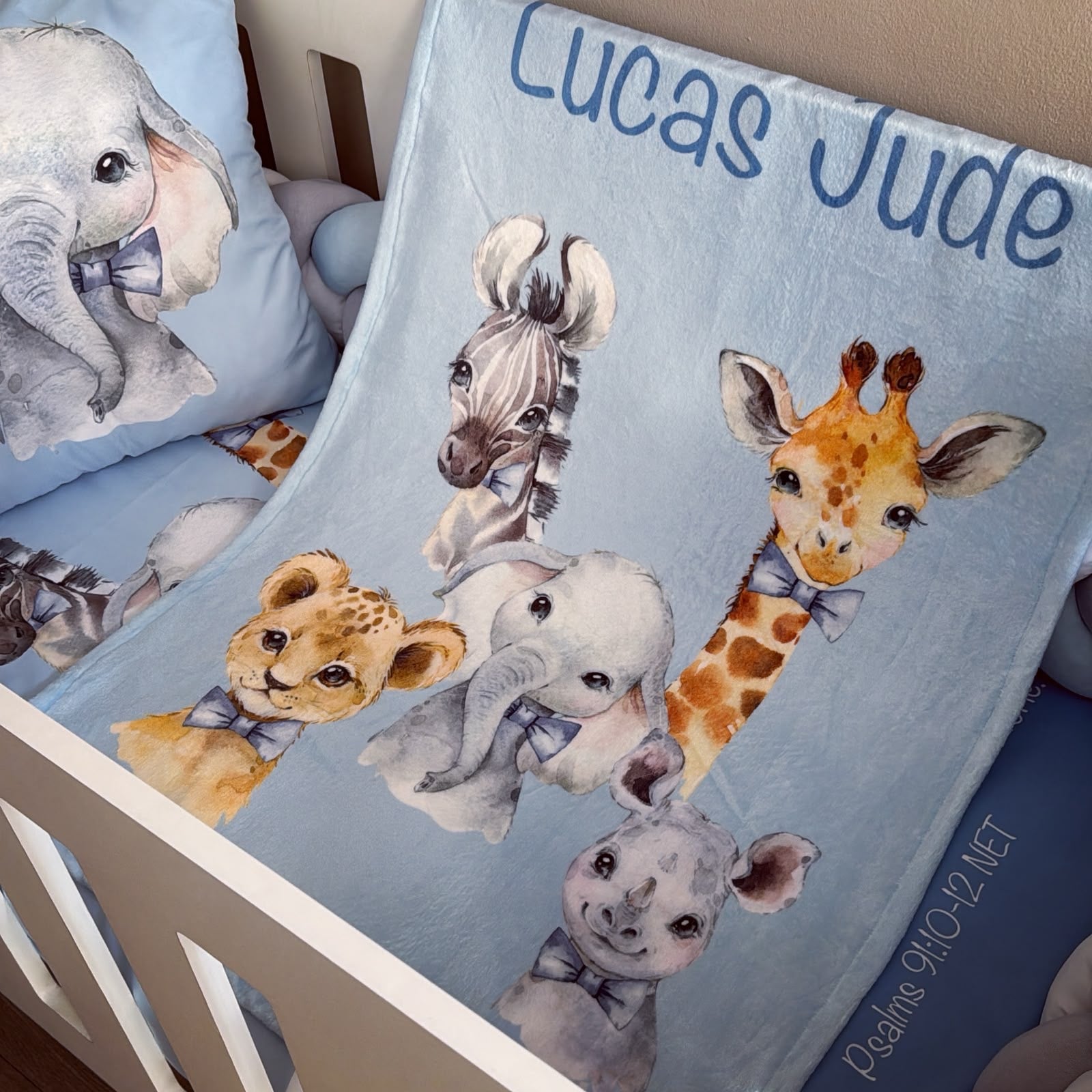 A personalized baby blanket draped over a white crib, featuring watercolor illustrations of an elephant, zebra, giraffe, lion cub, elephant, and rhino with bows, alongside the name 'Lucas Jude' in blue text.