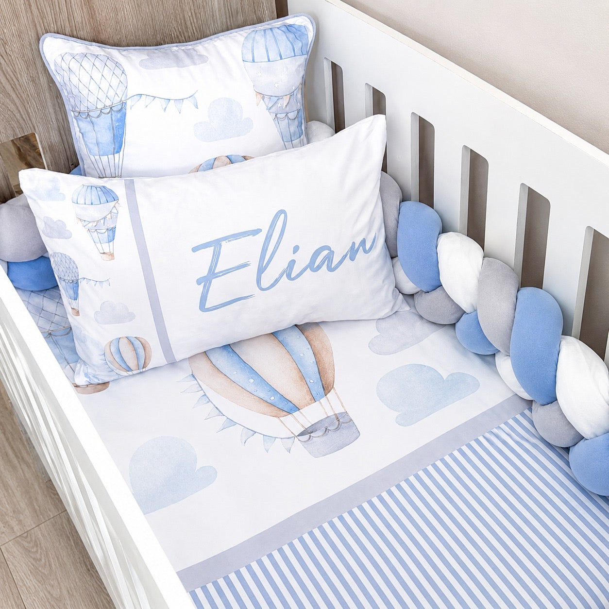 A baby bedding set styled in a white wooden crib, featuring a white sheet with blue stripes, a matching blue and white hot air balloon print, a personalized white pillow with the name 'Elian' in blue script, and coordinating pillows and a blue-and-white ball crib bumper.