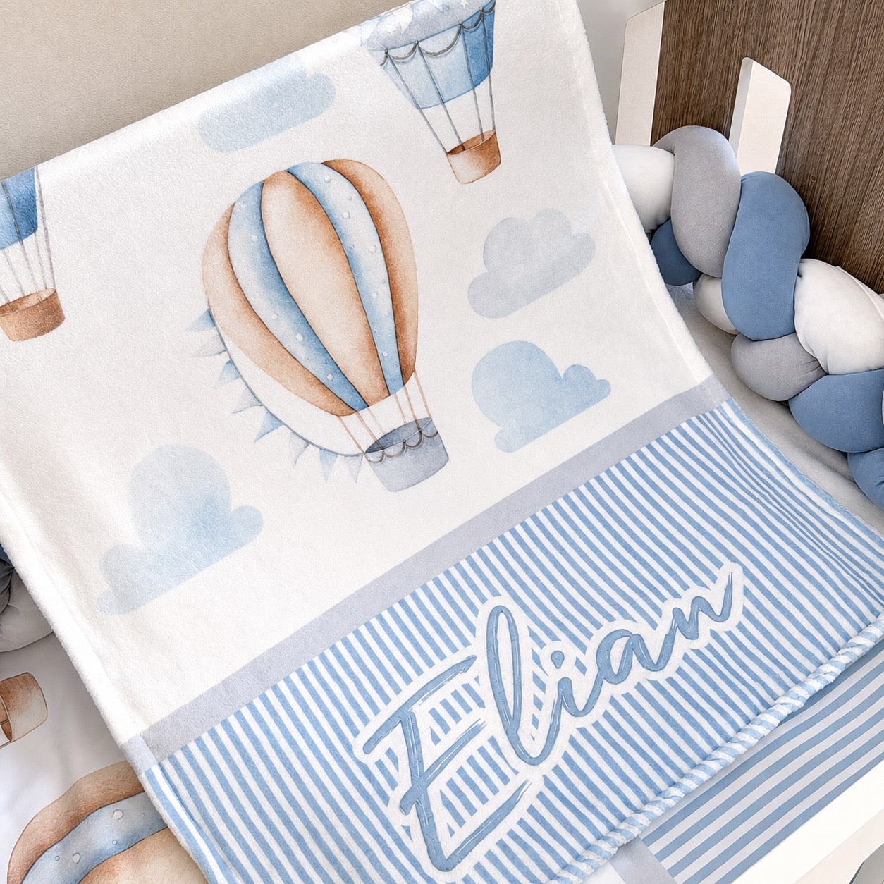 A personalized baby blanket with a white top section featuring watercolor hot air balloons and clouds, and a blue-and-white striped bottom section with the name 'Elian' in white script. The blanket is styled in a nursery setting next to a blue and white braided crib bumper.