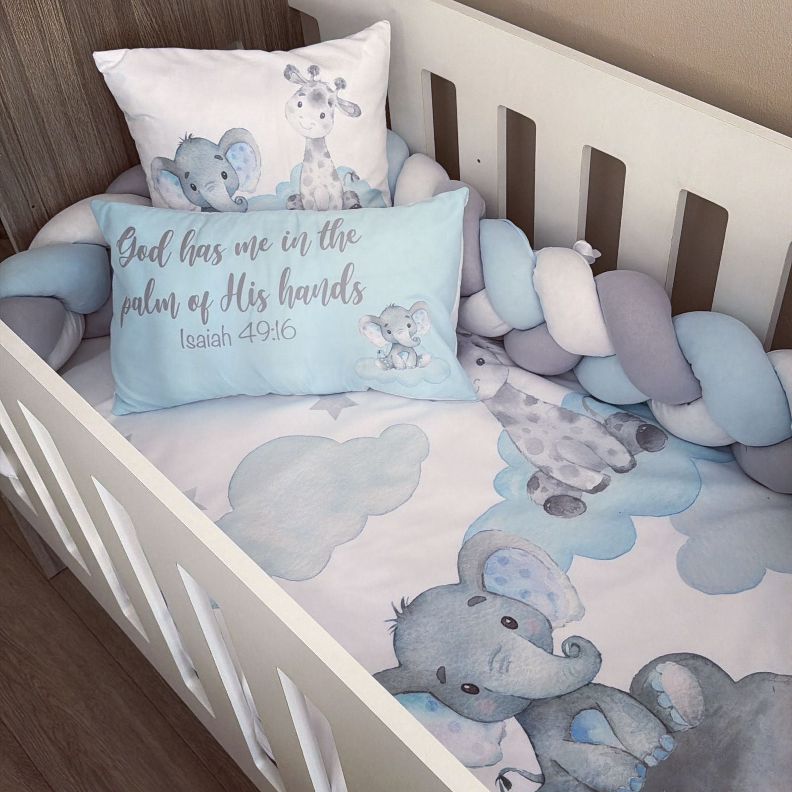 A baby crib bedding set featuring a light blue and gray elephant and giraffe print, a matching pillow with the text 'God has me in the palm of His hands Isaiah 49:16', and a chunky light blue, white, and gray bumper.