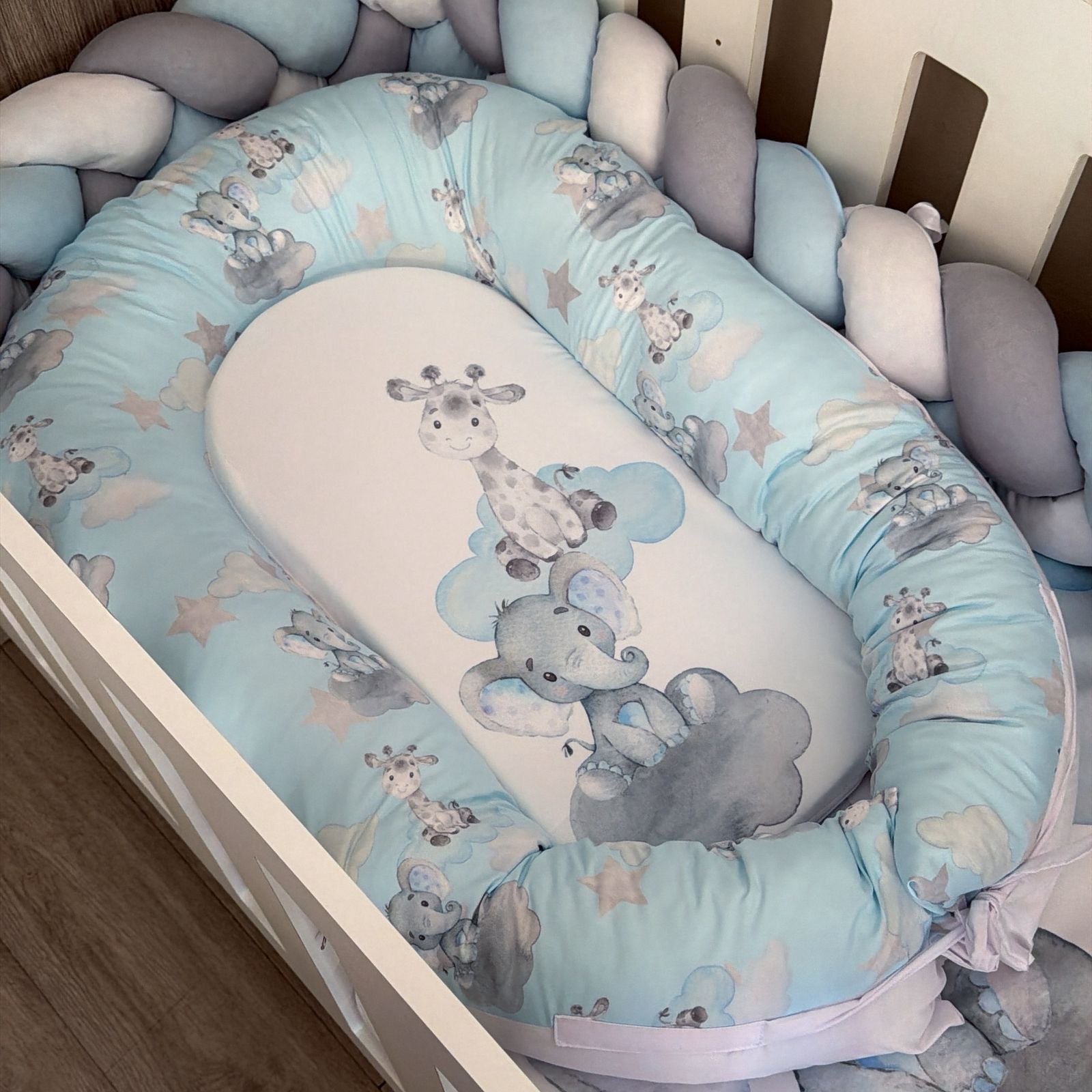 A baby bassinet bedding set with a light blue and white giraffe and elephant print, including a padded bassinet guard, mattress cover, and matching pillows inside a white crib.