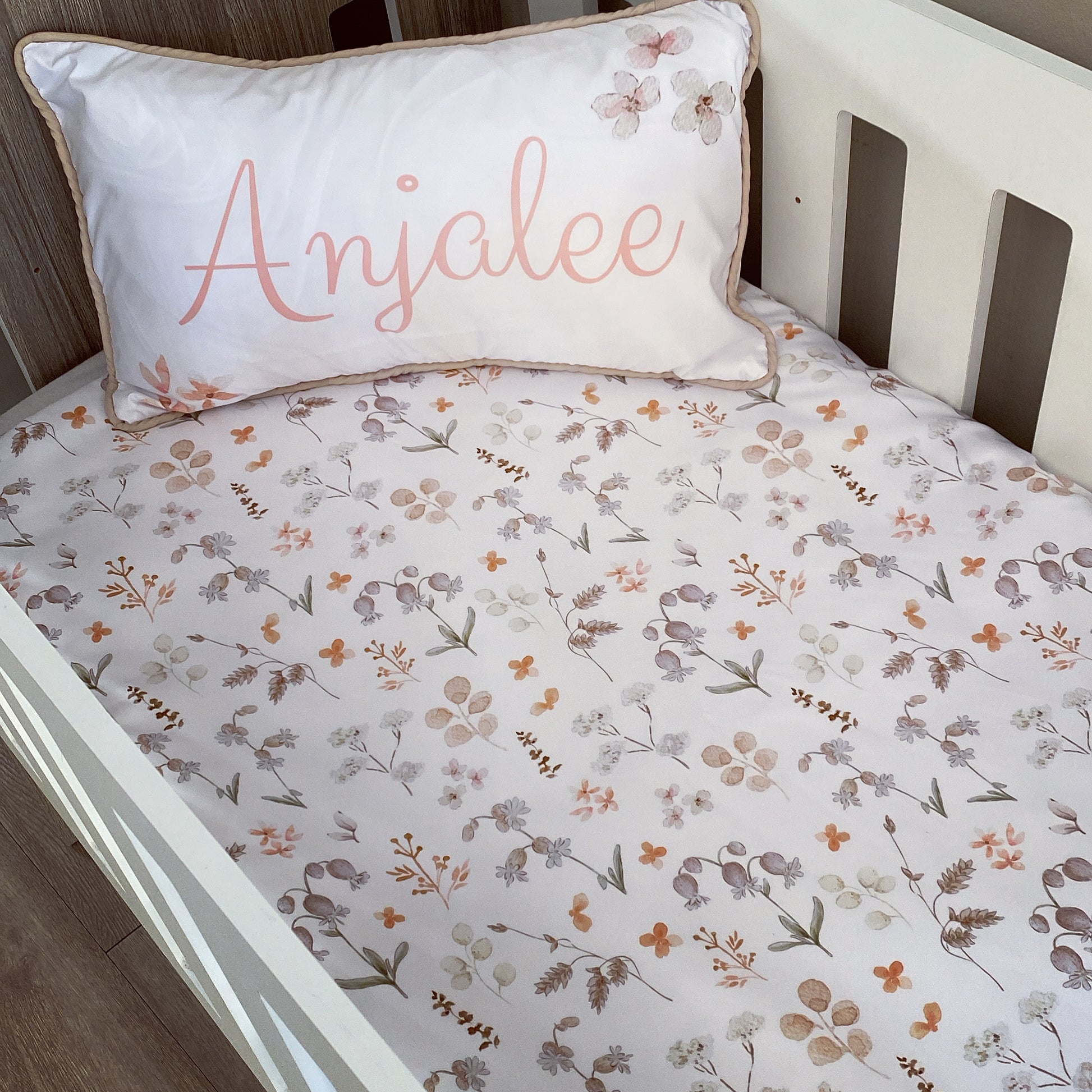 A cot with floral bedding and a pillowcase that has the name 'Anjalee' printed on it.