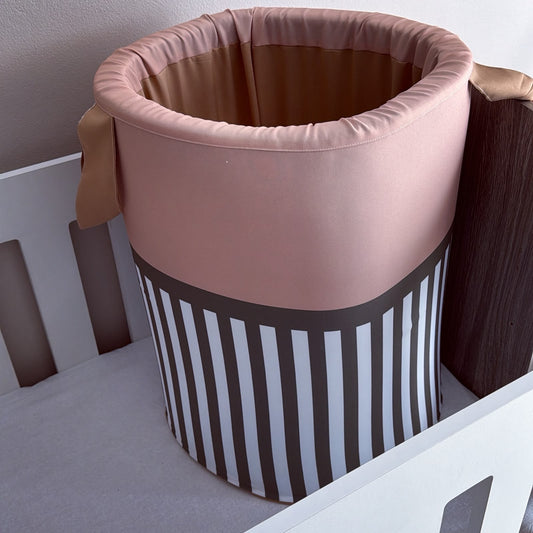 A fabric baby crib basket tan, and brown, featuring vertical brown and white stripes on the lower half, shown inside a white crib.
