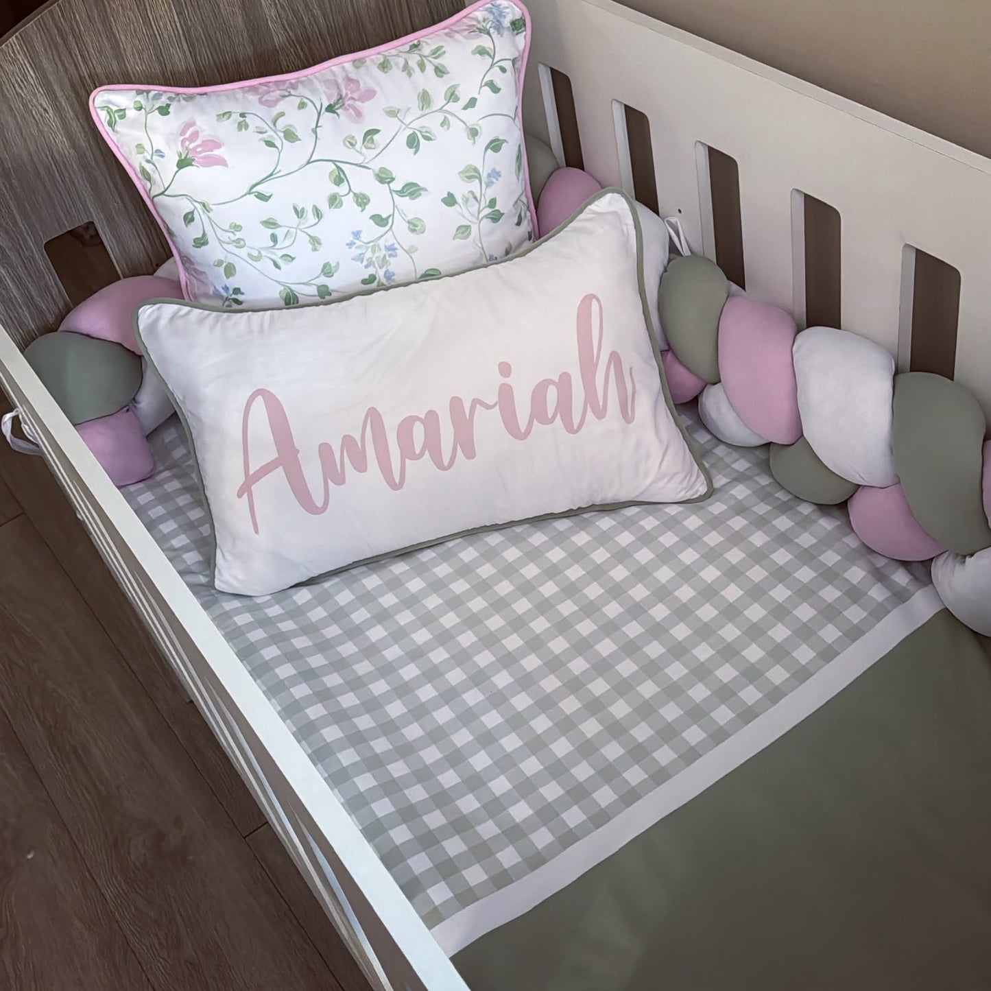 A styled baby crib setup featuring a personalized rectangular lumbar pillow with the name 'Amariah' in pink script, a gray and white gingham fitted sheet, a pink floral accent pillow, and a pastel chunky crib bumper.