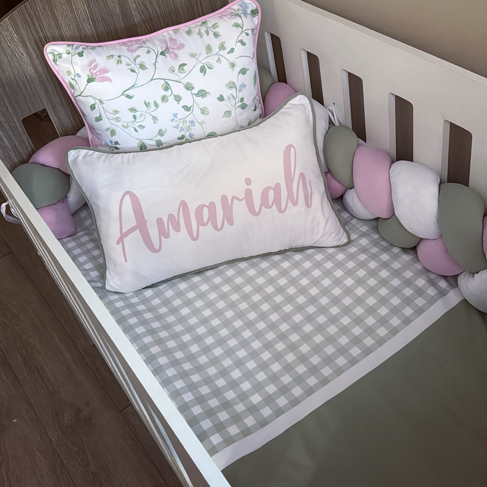 A styled baby crib setup featuring a personalized rectangular lumbar pillow with the name 'Amariah' in pink script, a gray and white gingham fitted sheet, a pink floral accent pillow, and a pastel chunky crib bumper.