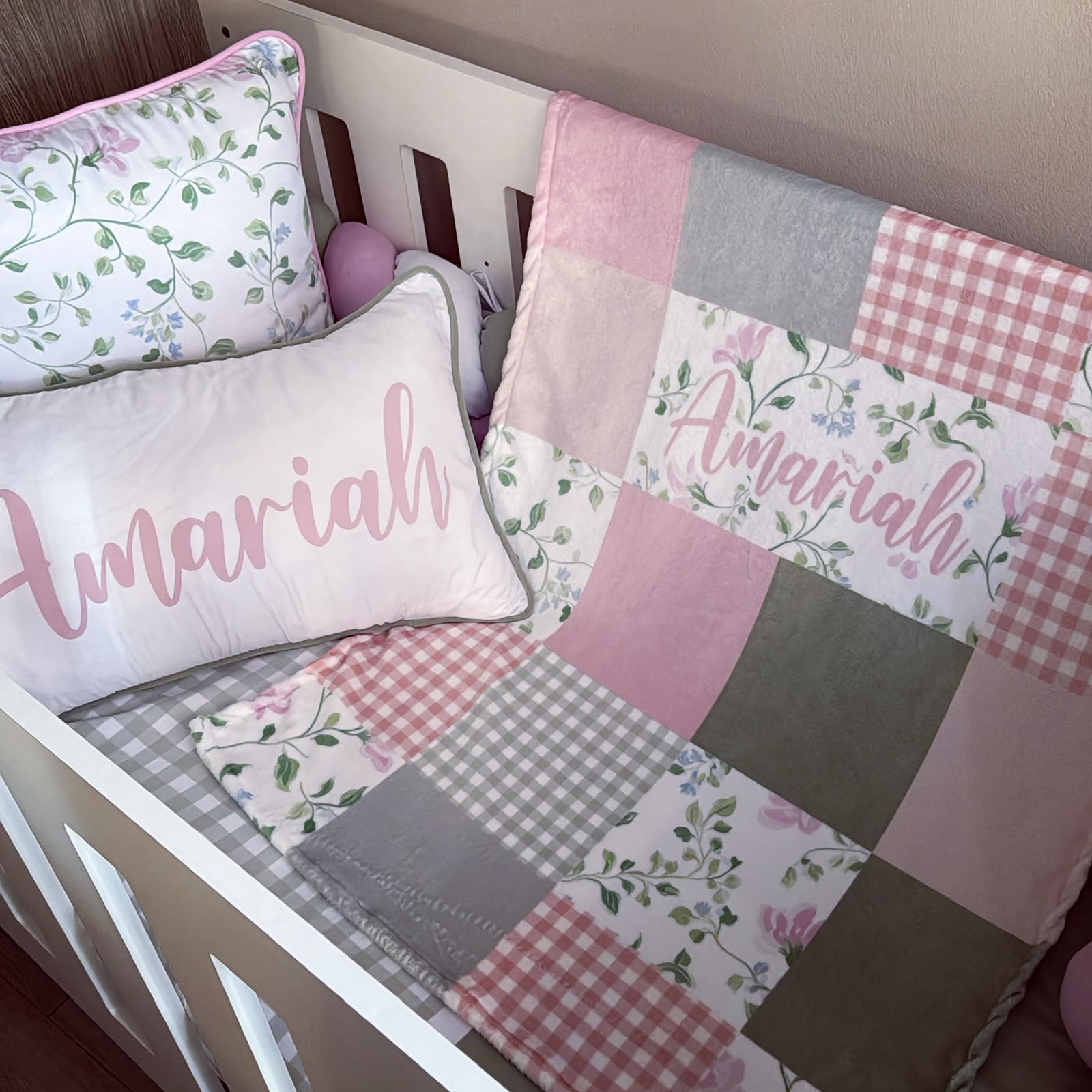A pink and white patchwork baby bedding set in a white crib, featuring a quilt with floral and gingham patterns, a matching pillow, and a personalized name pillow that reads 'Amariah'.