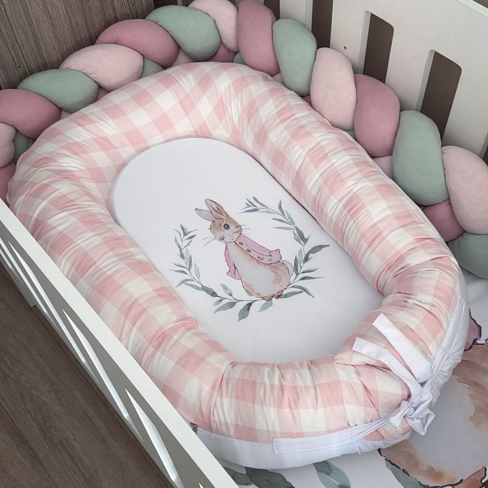 A baby bassinet cushion set in a white crib, featuring a pink and white gingham check padded bassinet pad with a white oval center printed with a watercolor bunny in a leafy wreath. The crib is surrounded by a braided bumper in shades of pink, sage green, and white.