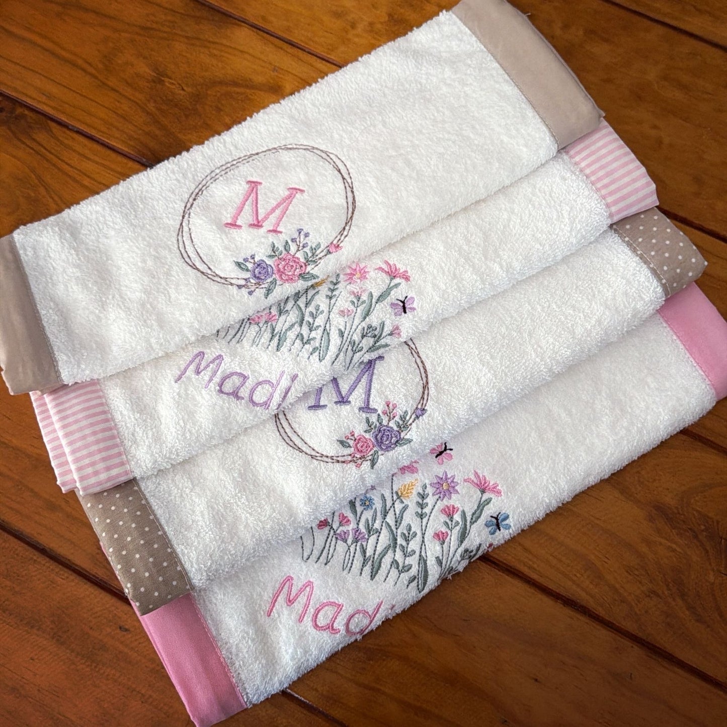 Three folded white burp cloths stacked on a wooden surface, each decorated with a floral monogram design and a different striped or polka-dot border trim.
