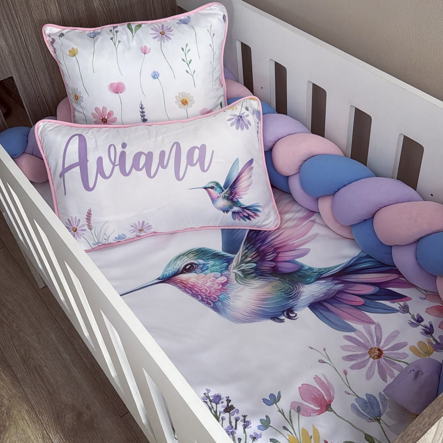 A personalized baby bedding set in a white wooden crib, featuring a printed sheet with a colorful hummingbird and floral motifs, a matching pillow with pink trim, and an additional rectangular pillow personalized with the name 'Aviana' in purple script.