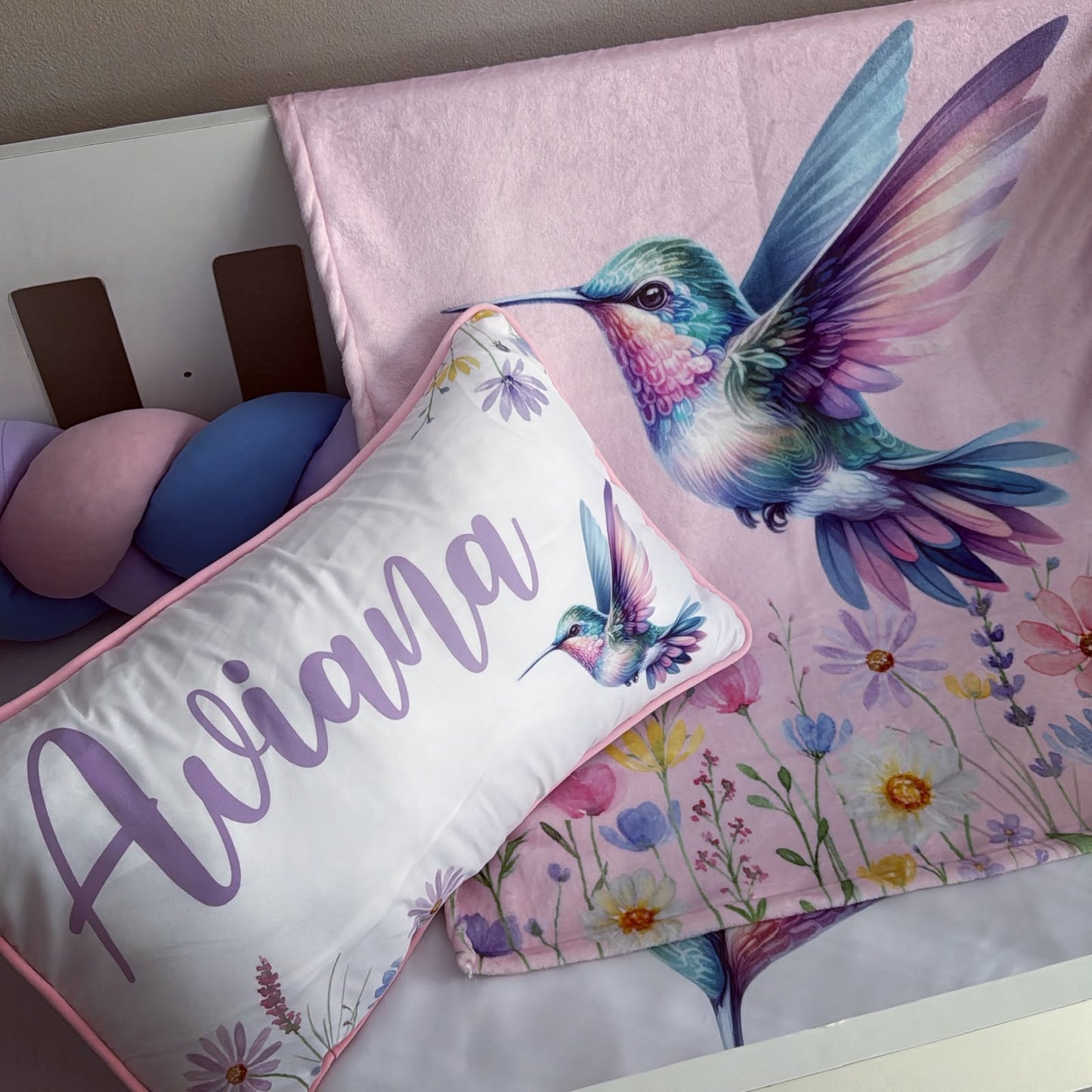 A pink baby blanket with a colorful hummingbird and floral print is styled next to a white personalized pillow with the name 'Aviana' in purple script.