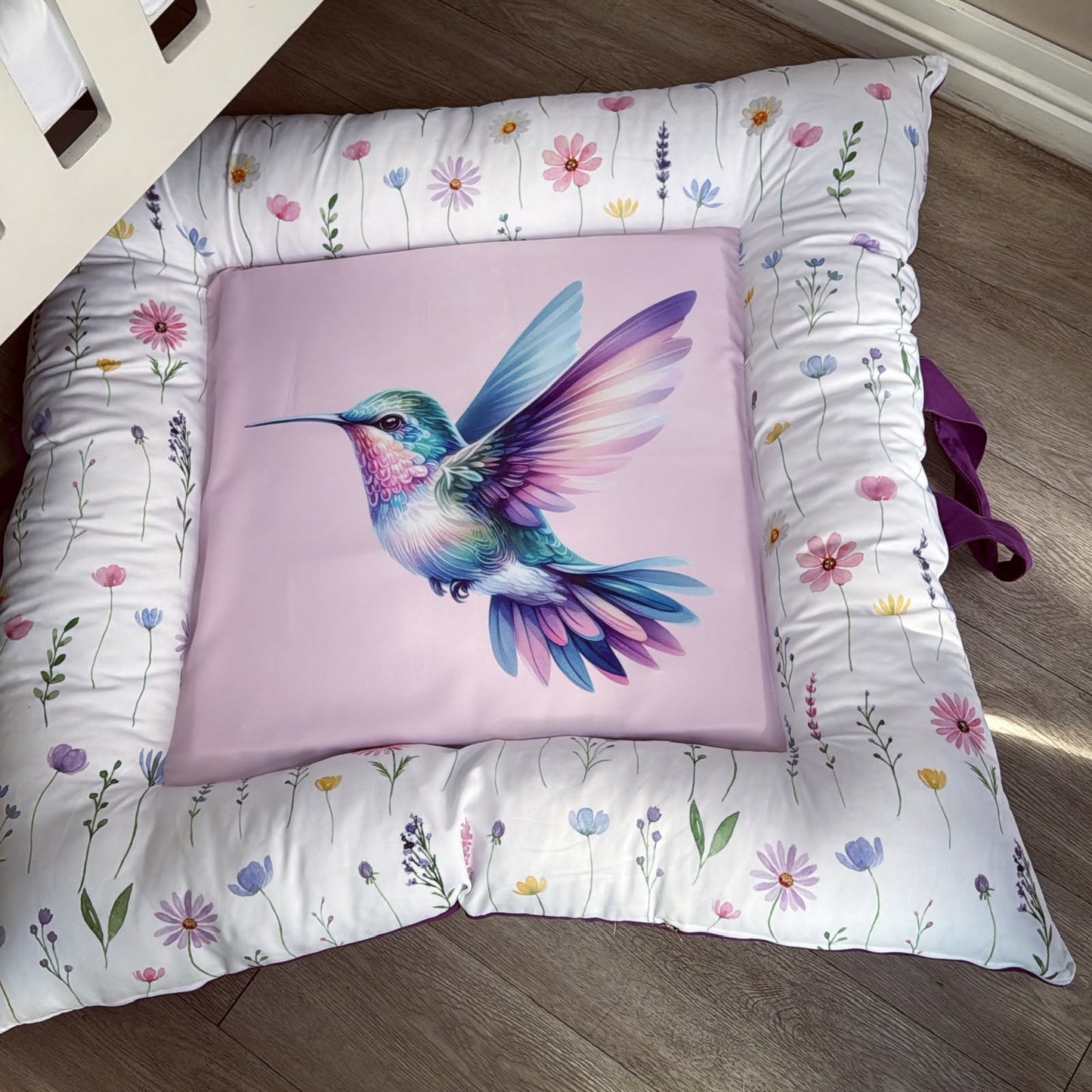 A square decorative cushion with a colorful hummingbird illustration on a light pink center panel, surrounded by a white floral border, photographed on a wooden floor.