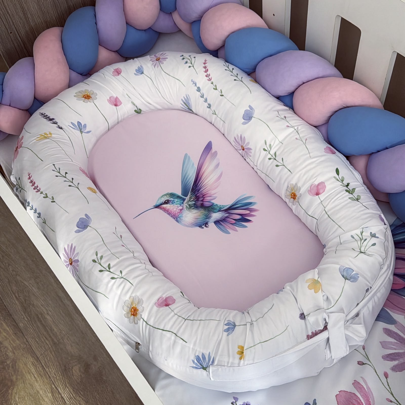 An oval baby bassinet with a white floral fabric lining and a pink insert cushion printed with a colorful hummingbird, styled inside a crib with a pastel bubble bumper.