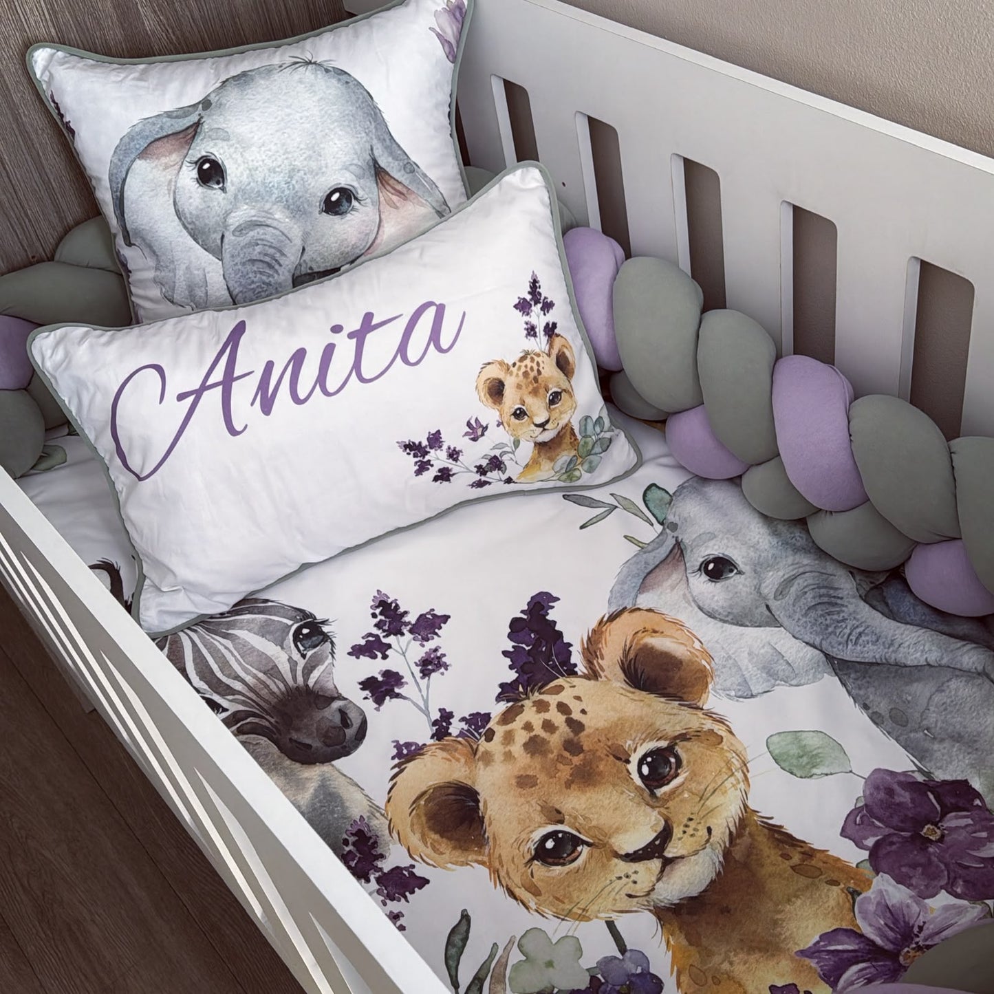 A personalized baby bedding set in a white crib, featuring a printed sheet and bumper with watercolor illustrations of a cheetah, elephant, zebra, and lavender flowers. Two matching pillows are included, one printed with a large elephant and the other personalized with the name 'Anita' in purple script alongside a small lion cub illustration.
