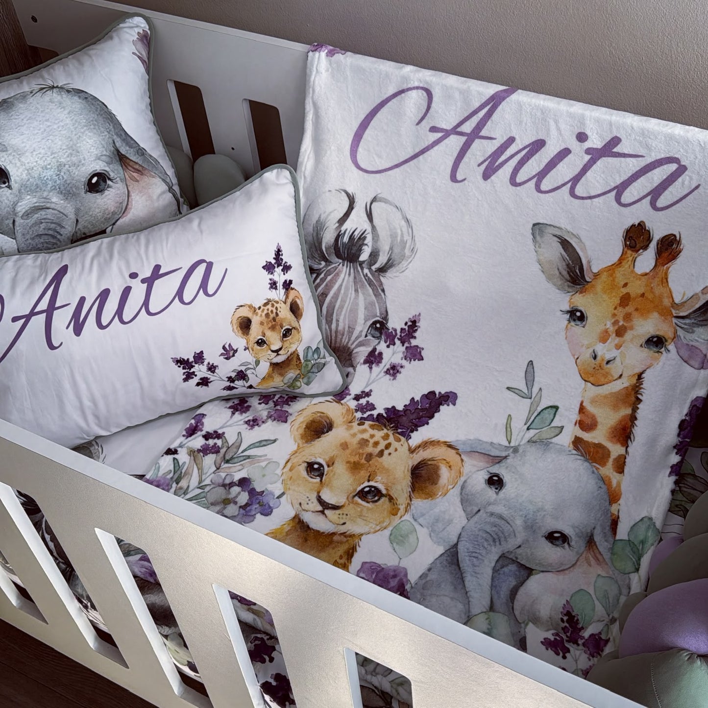 Custom safari-themed crib bedding set in a white wooden crib, featuring a duvet cover and matching pillowcases with watercolor illustrations of an elephant, giraffe, lion cub, and zebra, personalized with the name 'Anita' in purple script.